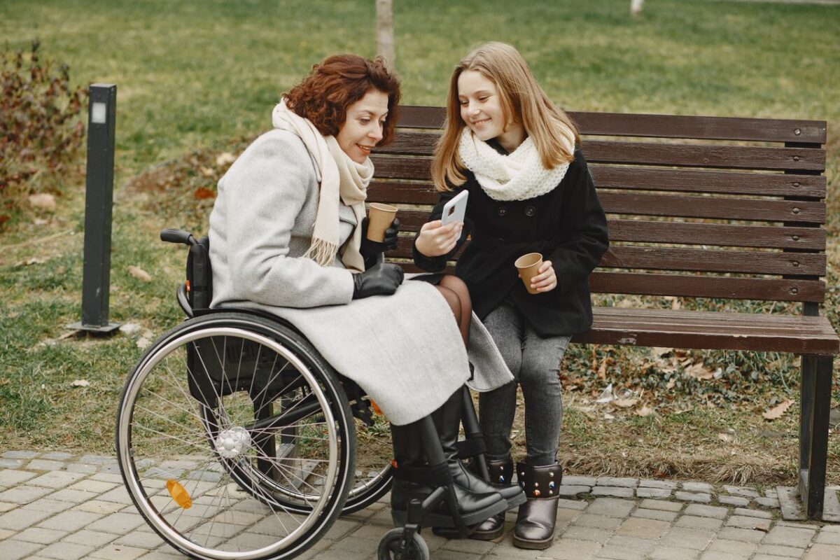 A woman in a wheelchair and her daughter sharing a joyful moment outdoors with a mobile phone.
